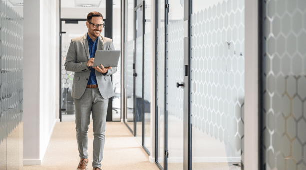 Professional man wearing glasses holding a laptop smiles and walks down hallway at work.