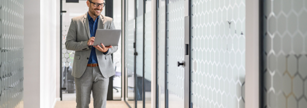 Professional man wearing glasses holding a laptop smiles and walks down hallway at work. 