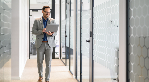 Professional man wearing glasses holding a laptop smiles and walks down hallway at work. 