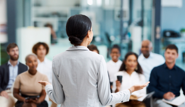 Back of woman as she speaks to a group of employees about benefits.